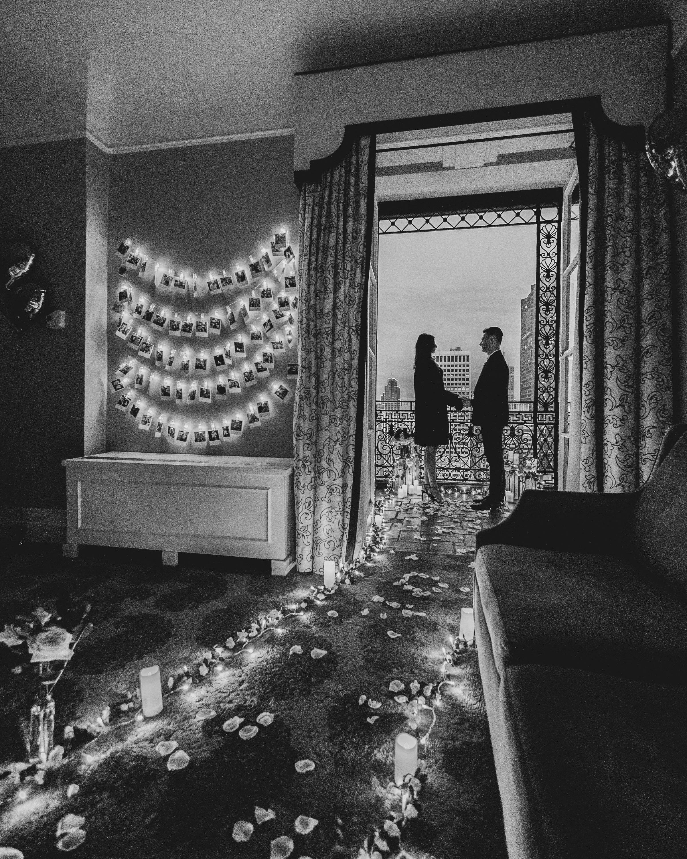 Black and white view from inside a Fairmont room showing candles, flower petals, string lights, and photos leading to the balcony proposal setup